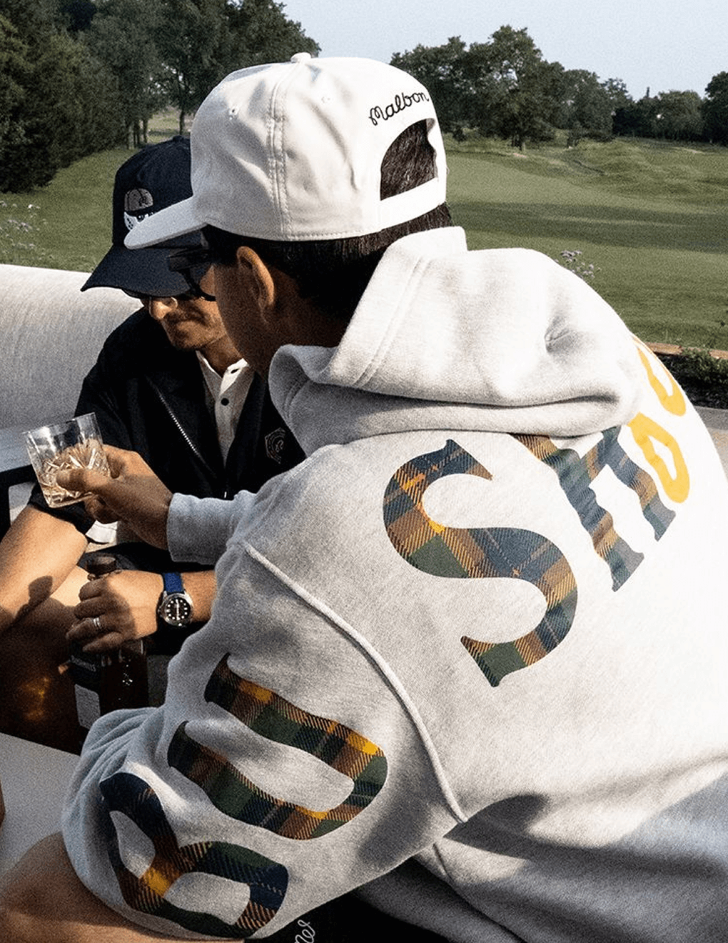 Detail shot of back of gray Bushmills x Malbon hoodie, man sitting with friends on golf course enjoying Bushmills.