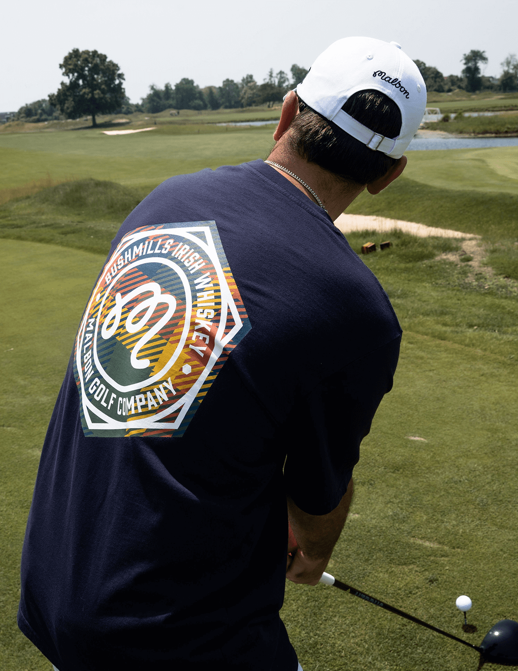 Back of navy Bushmills x Malbon tee worn by golfer swinging on golf course.