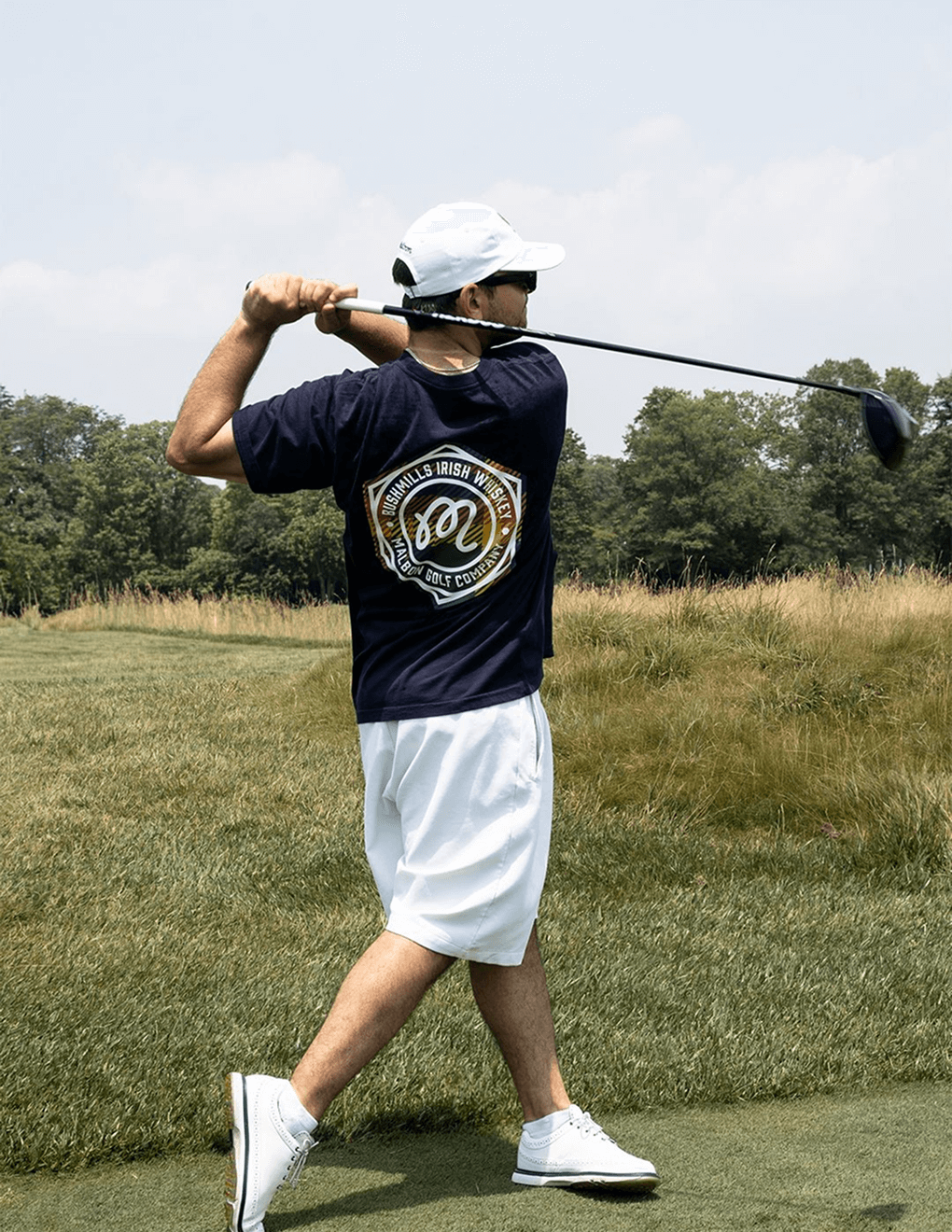 Navy Bushmills x Malbon tee worn by golfer swinging on golf course.