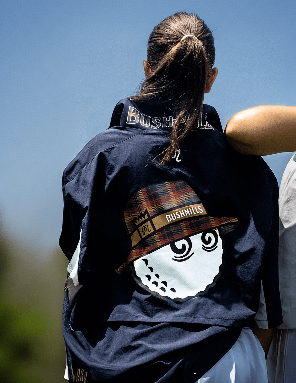 Woman wearing navy Bushmills x Malbon windbreaker, featuring back detail of Buckets cartoon.