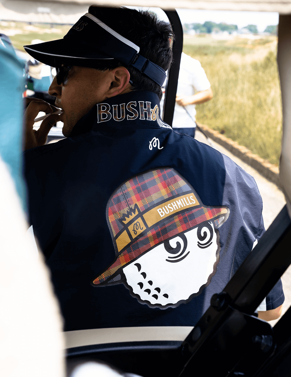 Man wearing navy Bushmills x Malbon windbreaker, featuring back detail of Buckets cartoon, while on a golf cart and wearing the Bushmills x Malbon visor.