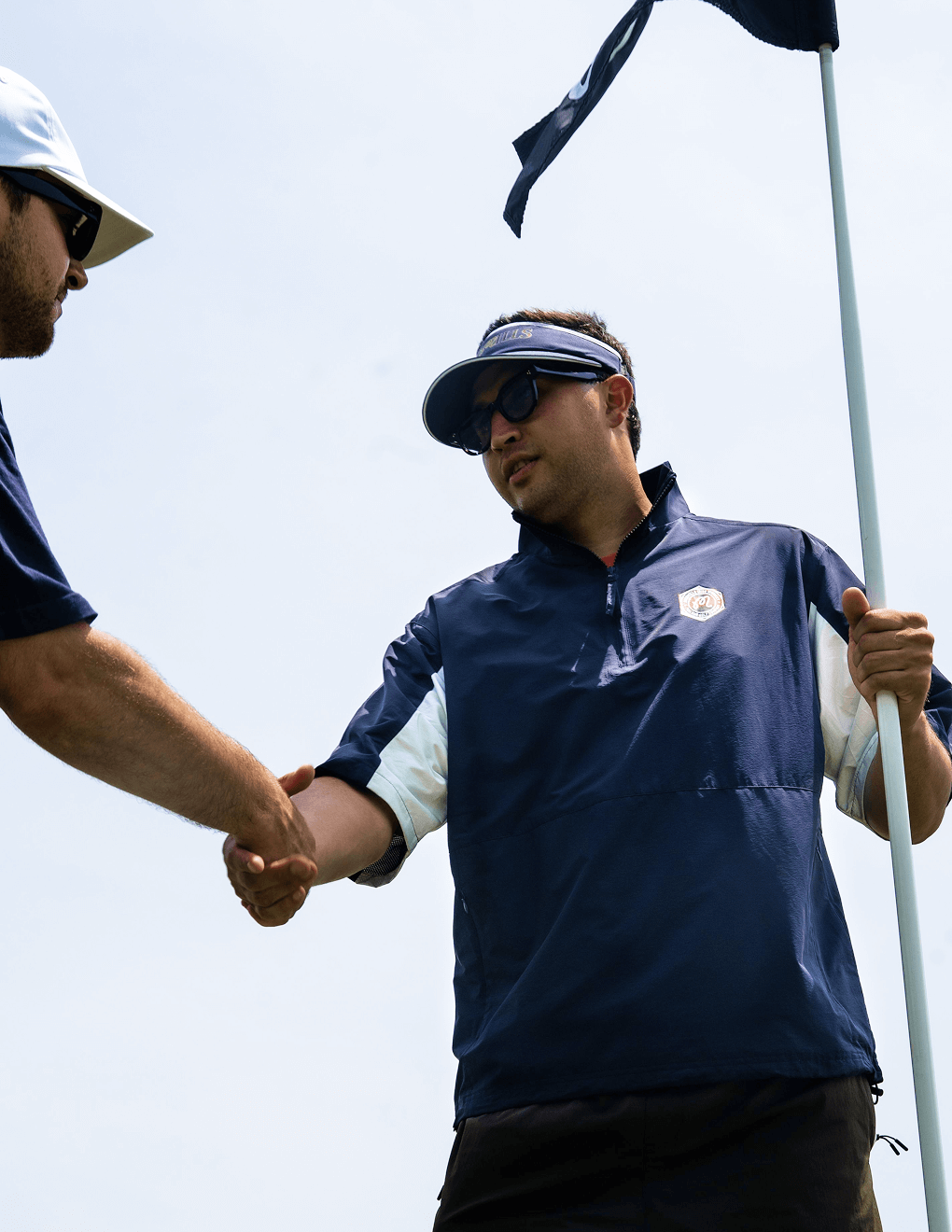 Man wearing navy Bushmills x Malbon windbreaker on the golf course, shaking hands with another golfer.