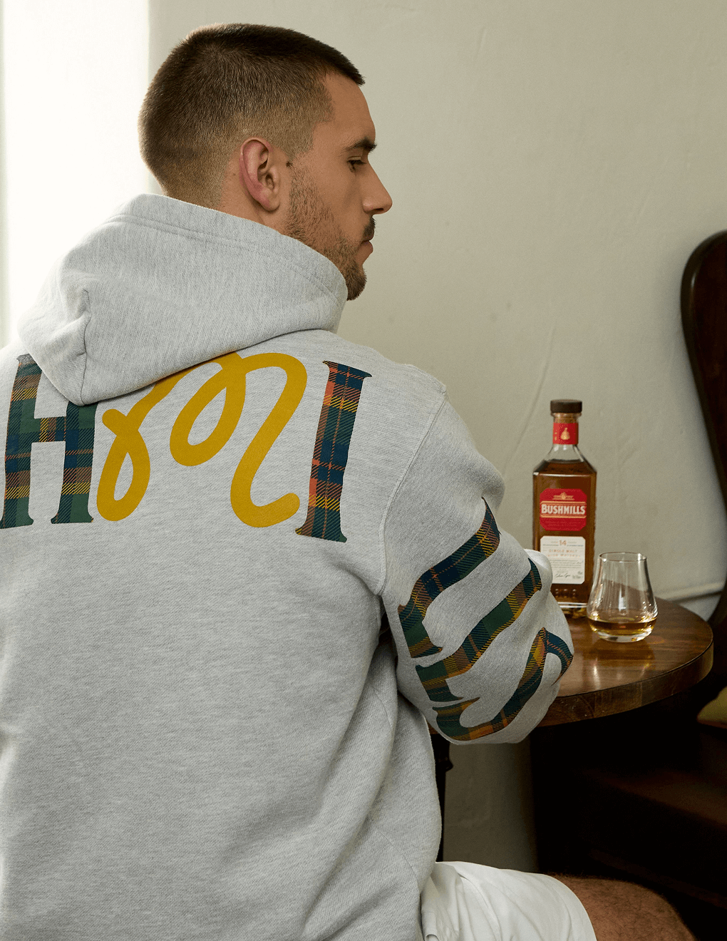 Detail shot of back of gray Bushmills x Malbon hoodie, man sitting in front of table with Bushmills 12 Year Single Malt.