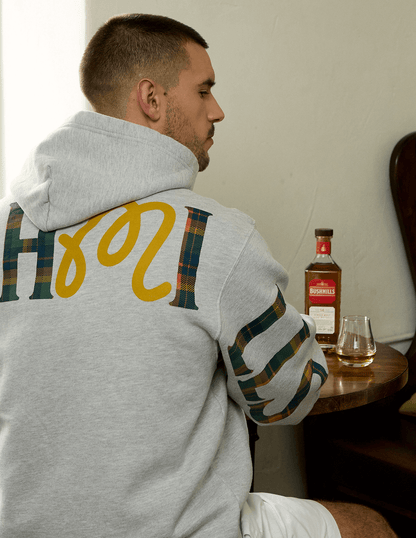 Detail shot of back of gray Bushmills x Malbon hoodie, man sitting in front of table with Bushmills 12 Year Single Malt.